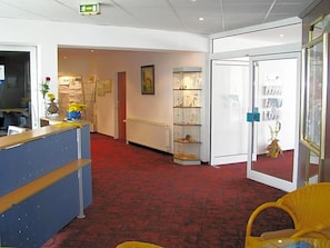 A hotel lobby with a reception desk, a red carpeted floor, a glass display case with items, and a door leading to another room.
