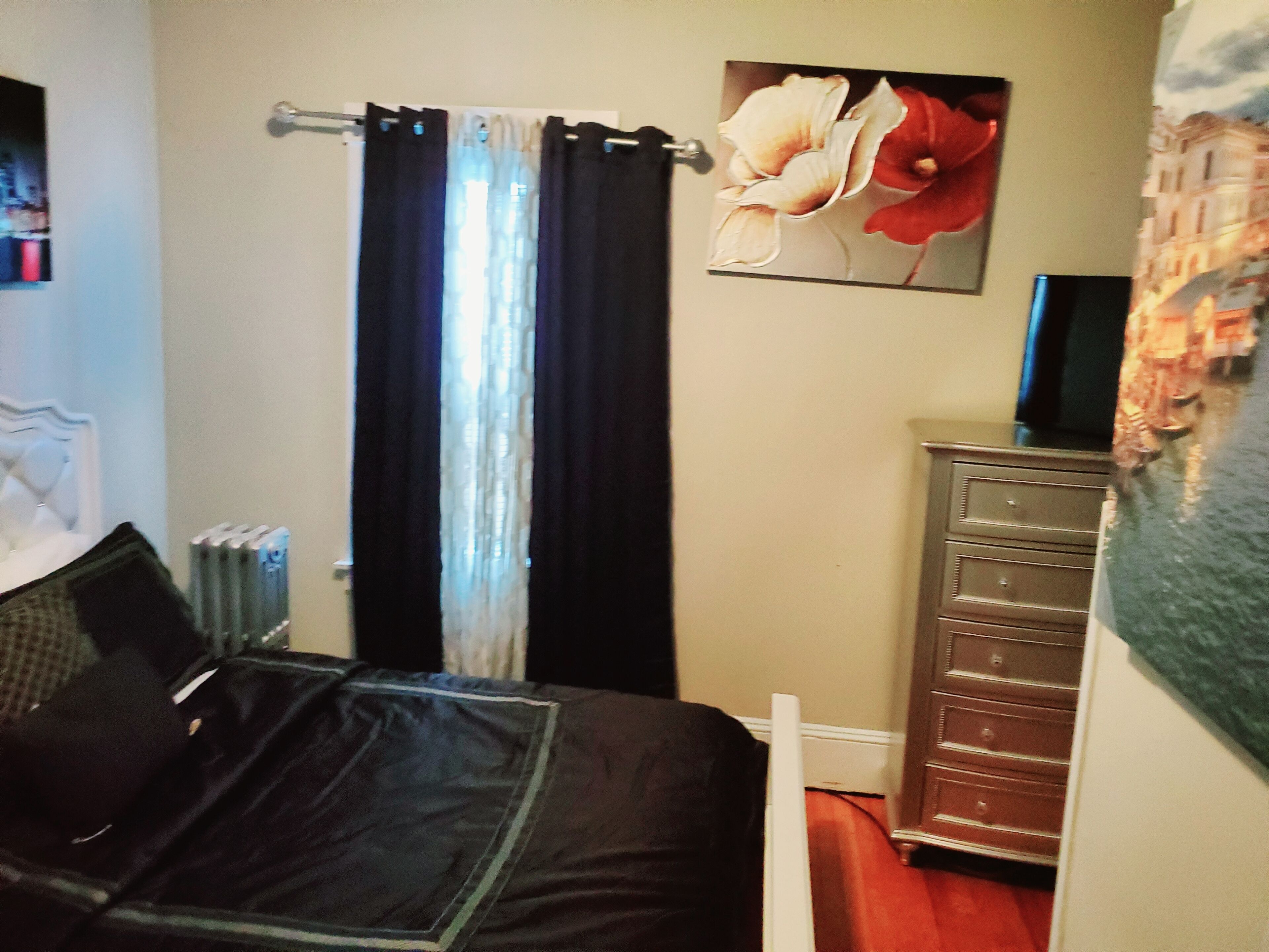 A bedroom with a bed, a radiator, a dresser, and a painting of flowers on the wall.