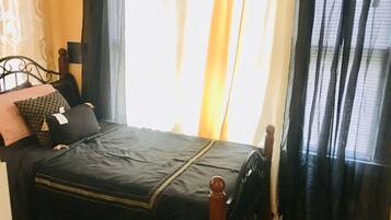 Ein Bett mit dunkler Bettdecke und einem hölzernen Kopfteil, ein Nachttisch mit Lampe sowie ein Fenster mit transparenten Vorhängen.