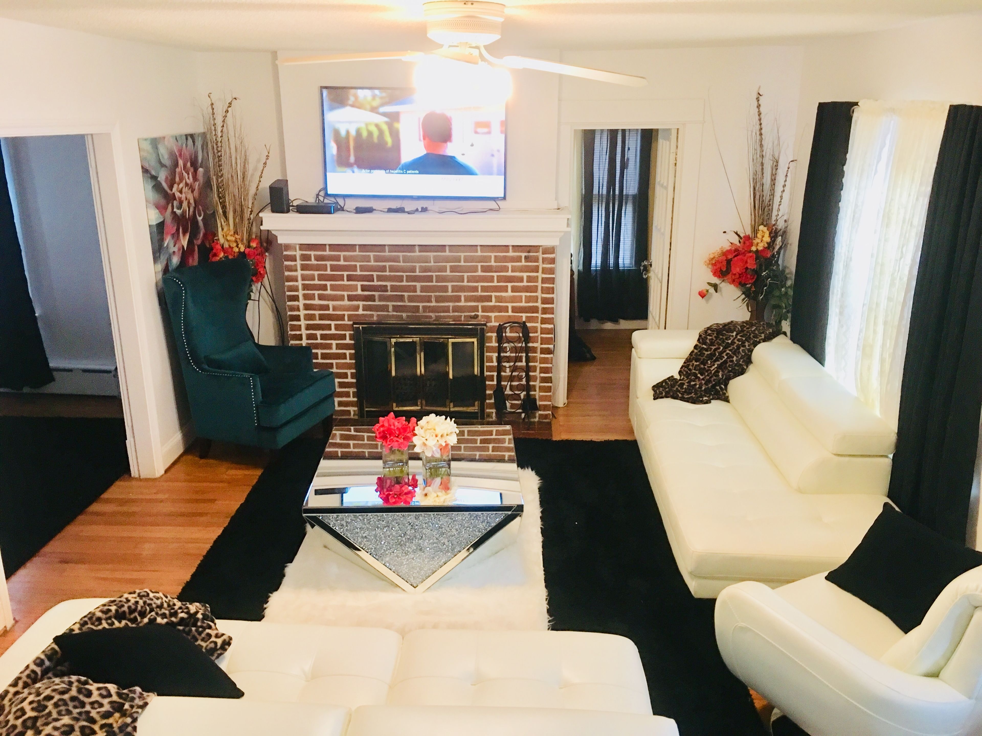 A living room with a brick fireplace, a television mounted above it, a white sofa, a teal armchair, and a coffee table with flowers.