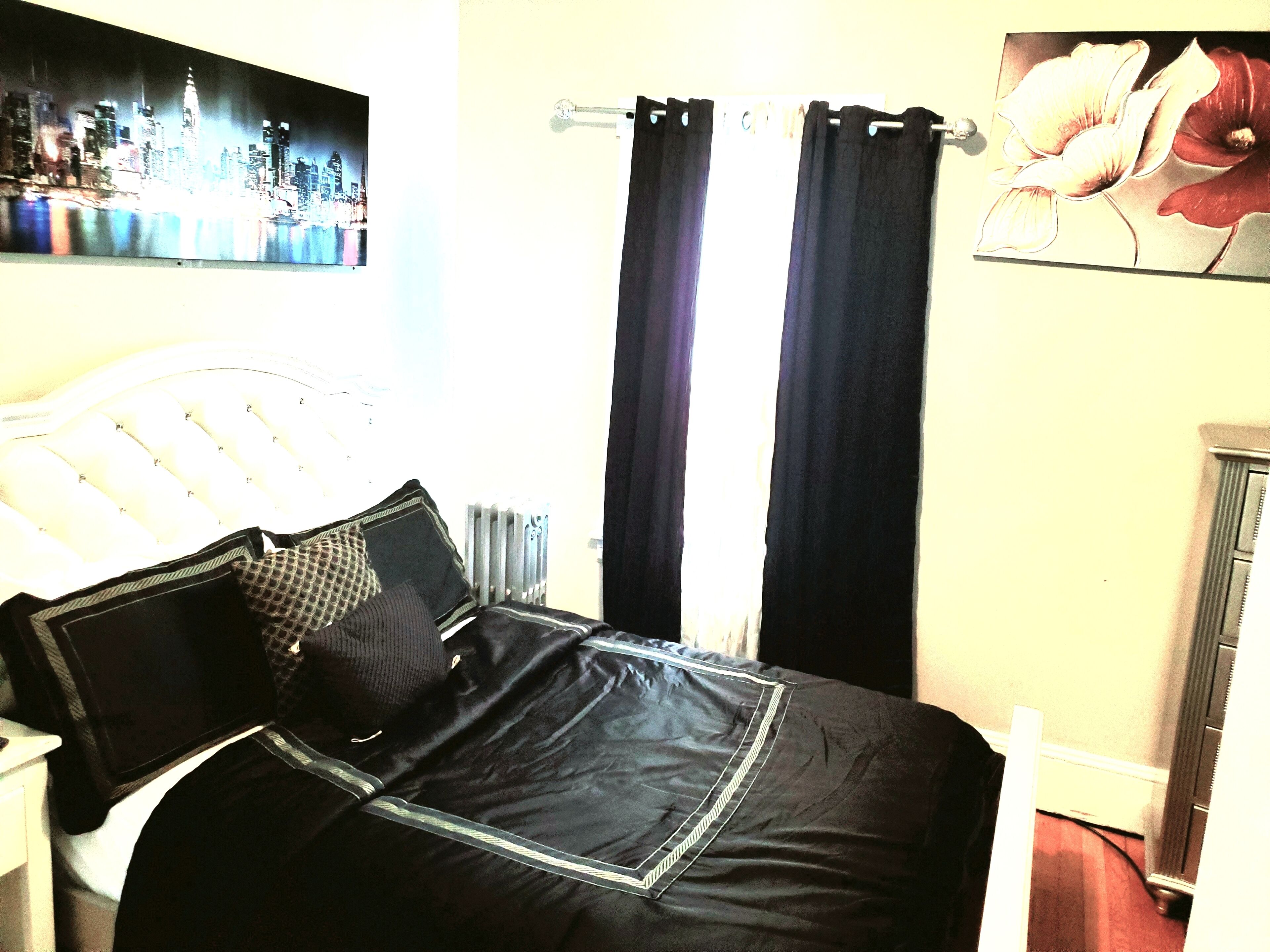 A bedroom with a bed, black curtains, a radiator, and a cityscape painting on the wall.