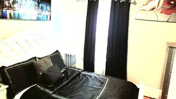 A bedroom with a bed, black curtains, a radiator, and a cityscape painting on the wall.