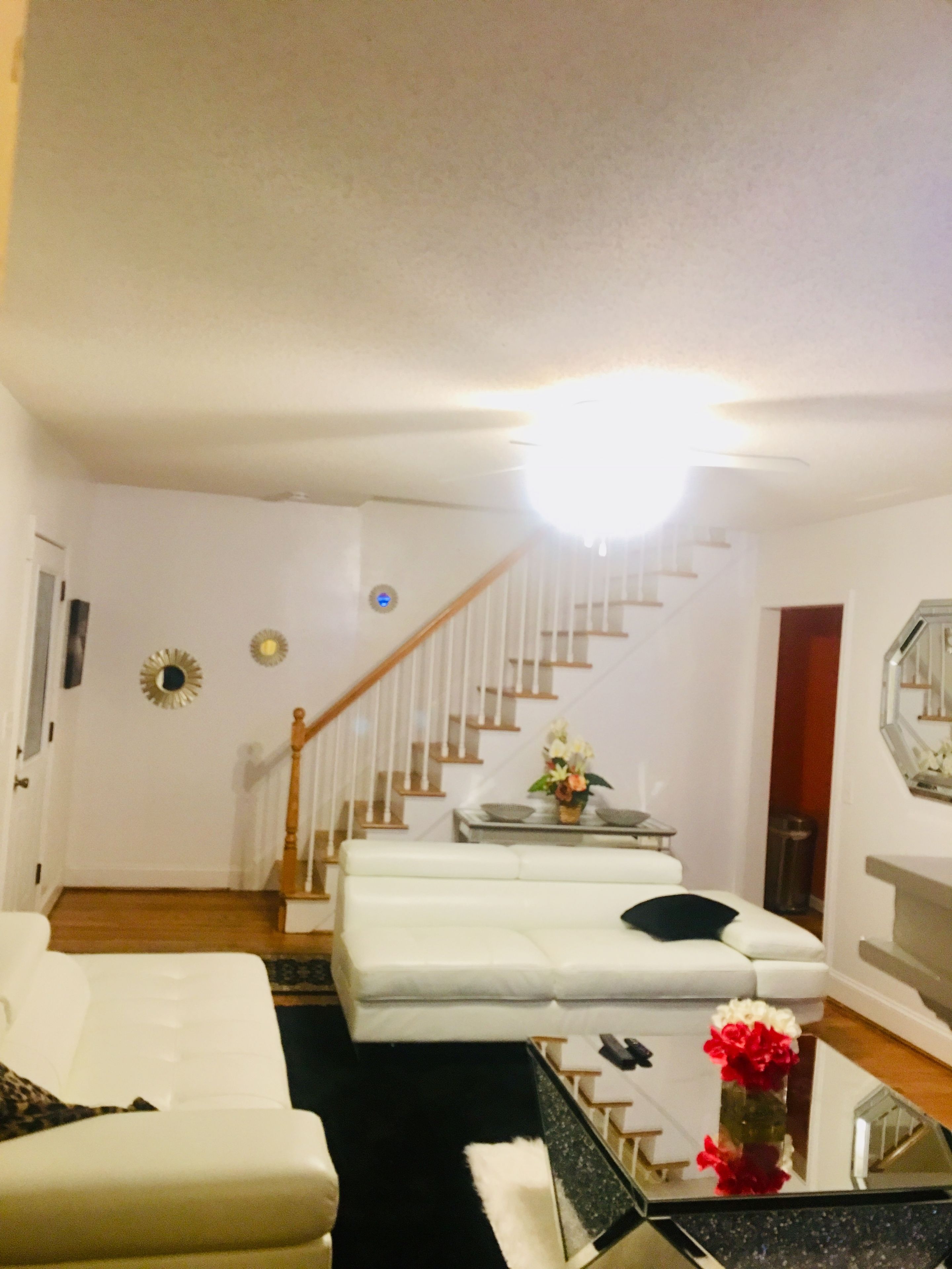A living room with a white sofa, a coffee table with a vase of flowers, a staircase with a wooden railing, and a white wall with a decorative mirror.