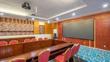 A conference room with a long table, red chairs, a projector screen, and a patterned carpet.