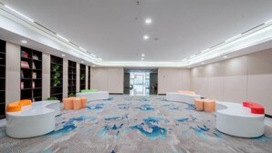 Eine geräumige Lobby mit modernem Design, mit einer zentralen Sitzecke mit weißen und orangefarbenen Stühlen, einem gemusterten Teppich und einer Bücherwand voller Bücher und Pflanzen auf der linken Seite.