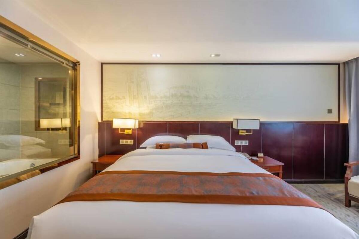 A hotel room with a large bed, a nightstand, a lamp, and a view of a building outside.