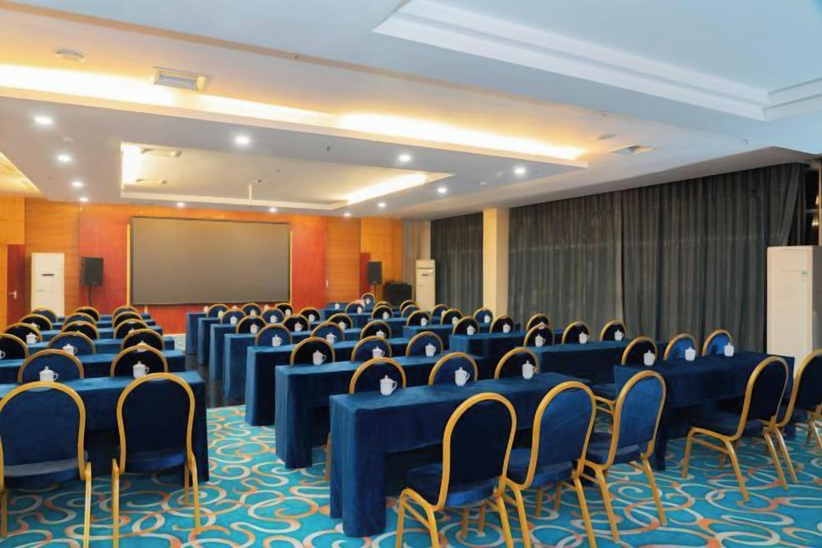 A conference room with rows of tables and chairs, a projector screen, and a stage.