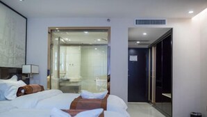 Ein Hotelzimmer mit Bett, einem durch eine Glastür einsehbaren Badezimmer und einem zur nächsten Zimmer führenden Gang.
