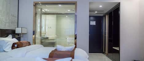 A hotel room with a bed, a bathroom visible through a glass door, and a hallway leading to another room.