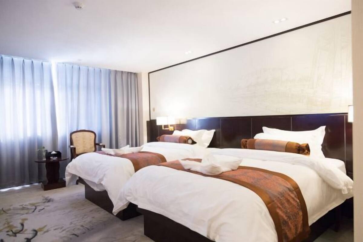 A hotel room with two beds, a large window with curtains, a small table, and a wall-mounted TV.