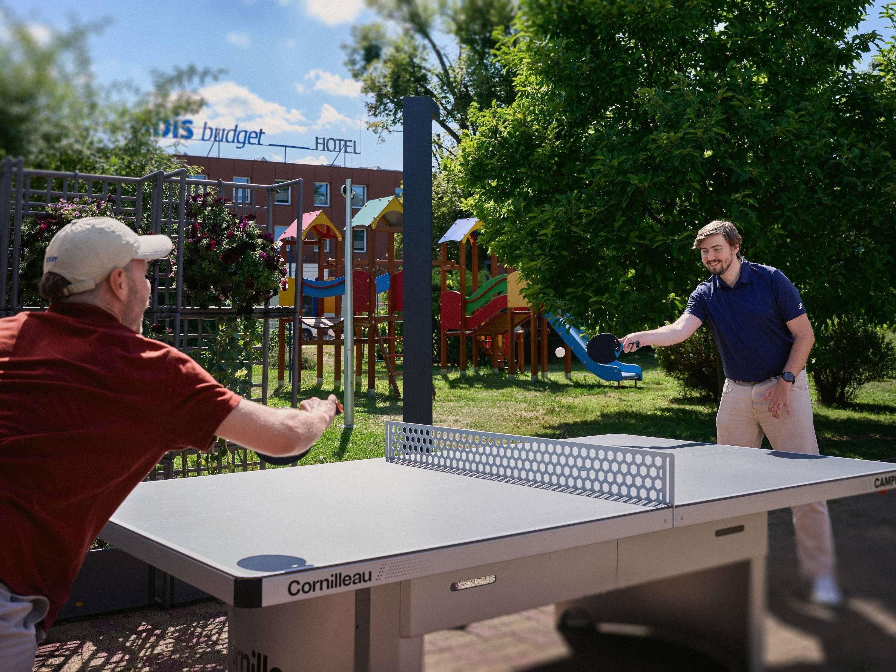 Dos personas jugando ping pong al aire libre, con un parque infantil y un hotel al fondo.
