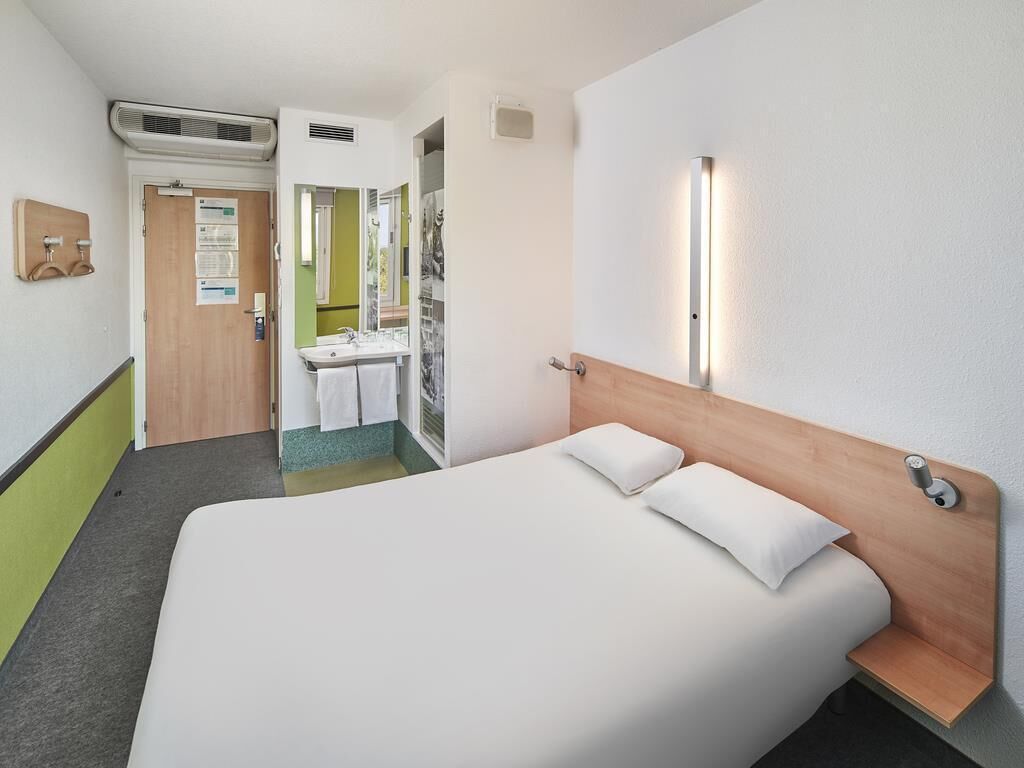 Quarto de hotel com cama, mesa de cabeceira, espelho, luminária na parede e porta para o banheiro.