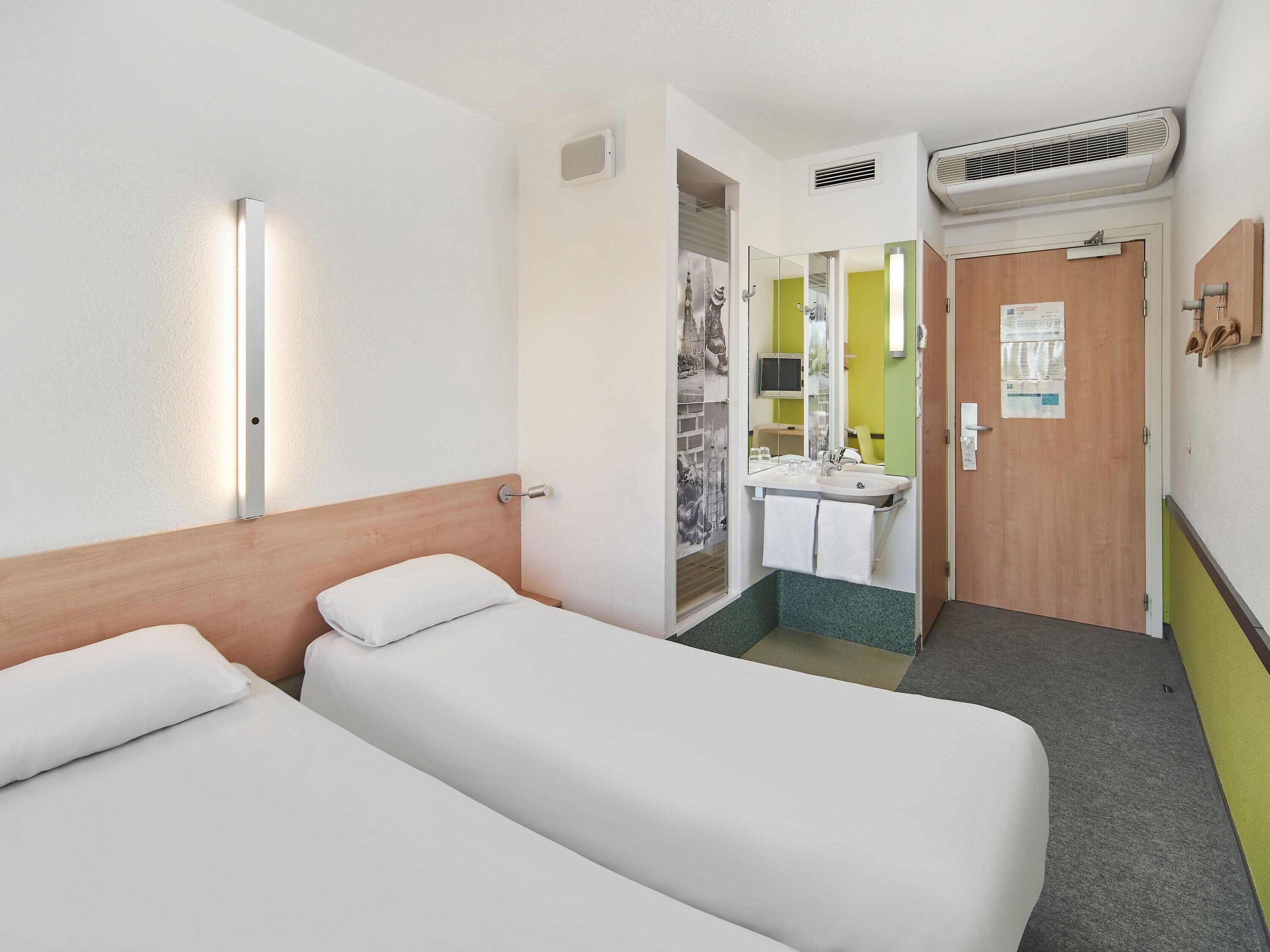 Habitación de hotel con dos camas individuales, baño con lavamanos y espejo, y pasillo con una puerta.