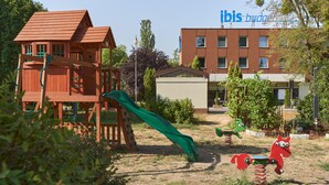 Un parque infantil con tobogán y columpios, rodeado de árboles y un edificio con el rótulo "ibis budget".