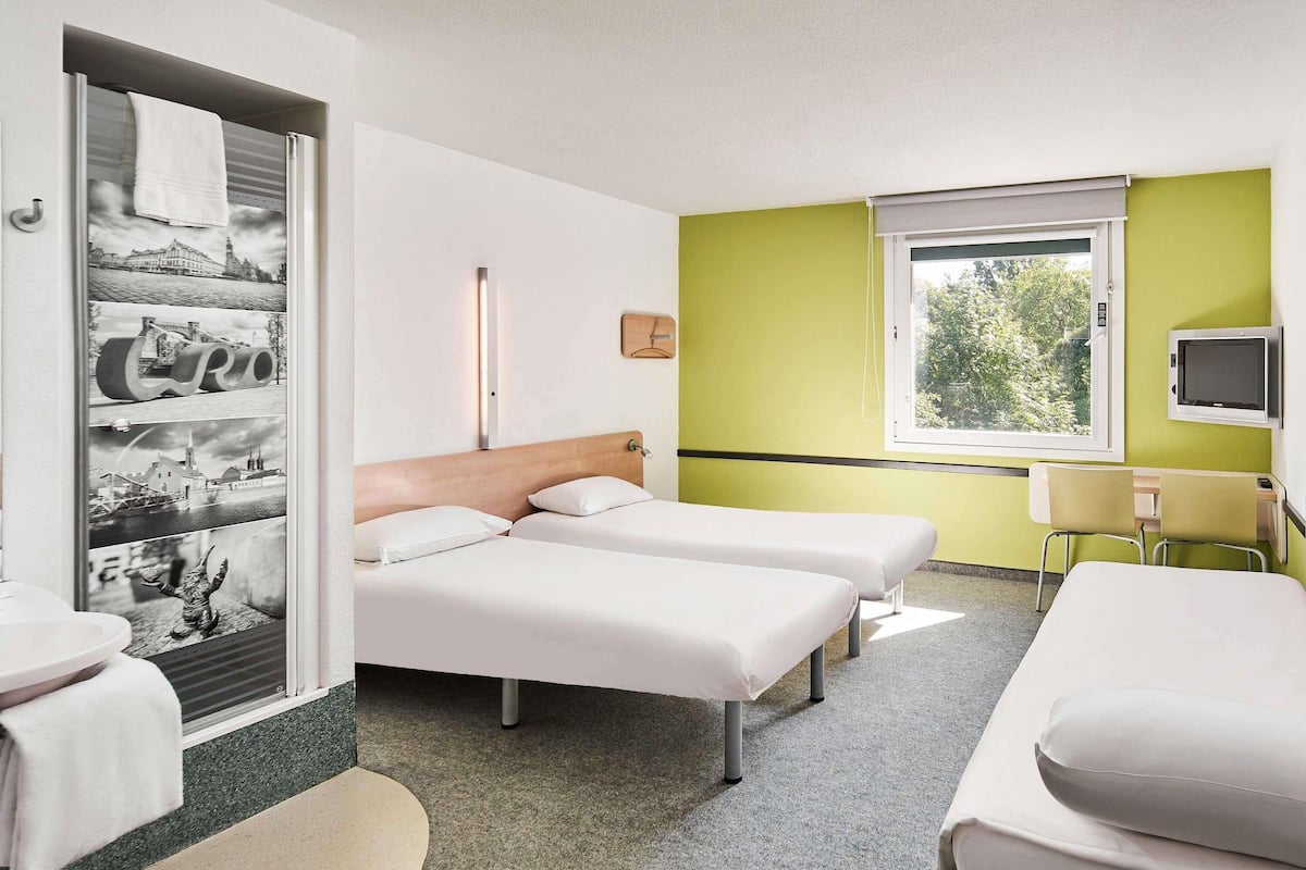 A hotel room with two beds, a desk, a chair, a television, and a window with a view of trees.