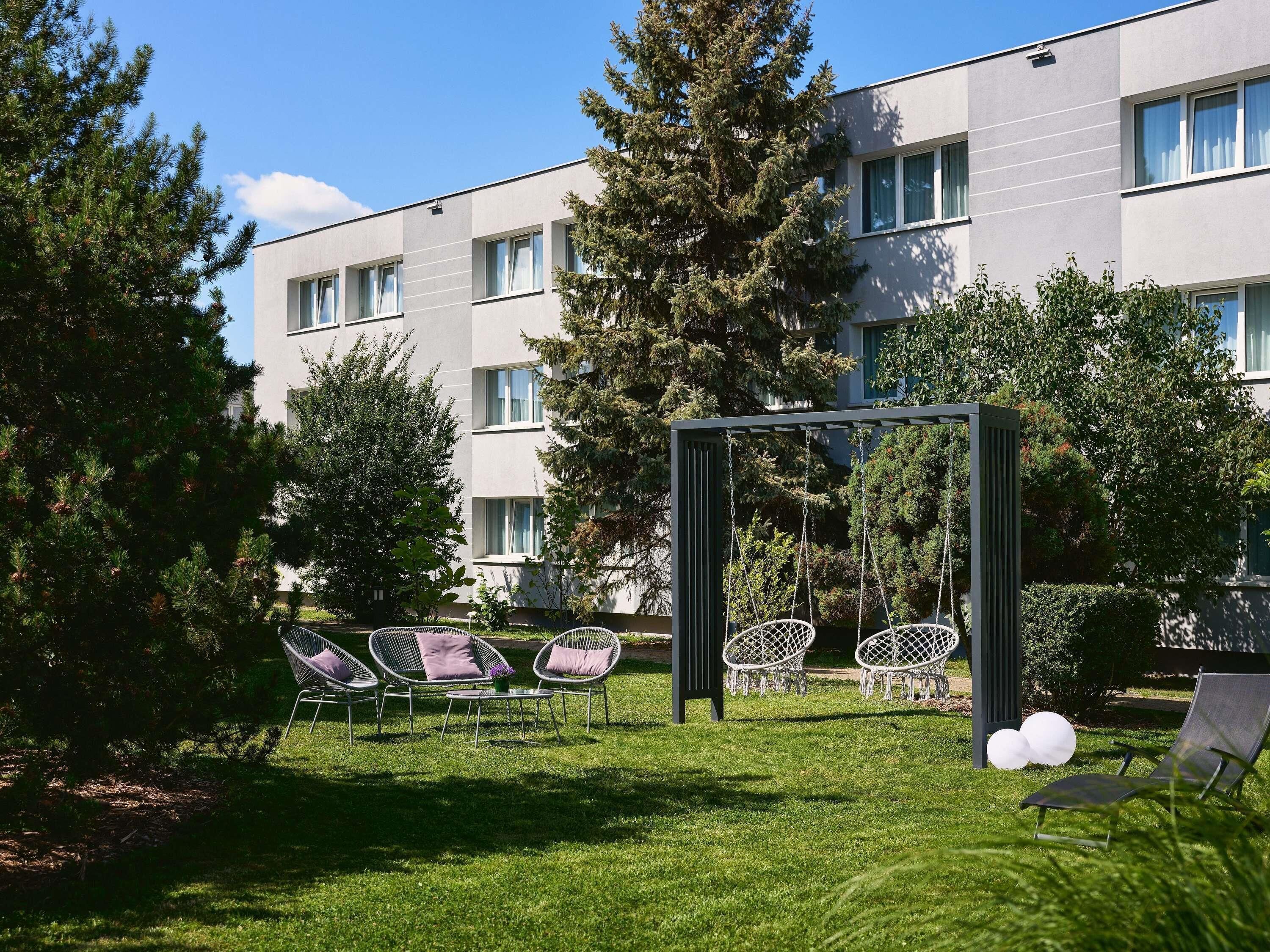 A modern outdoor seating area with chairs and a swing set, surrounded by greenery and a building with multiple windows.