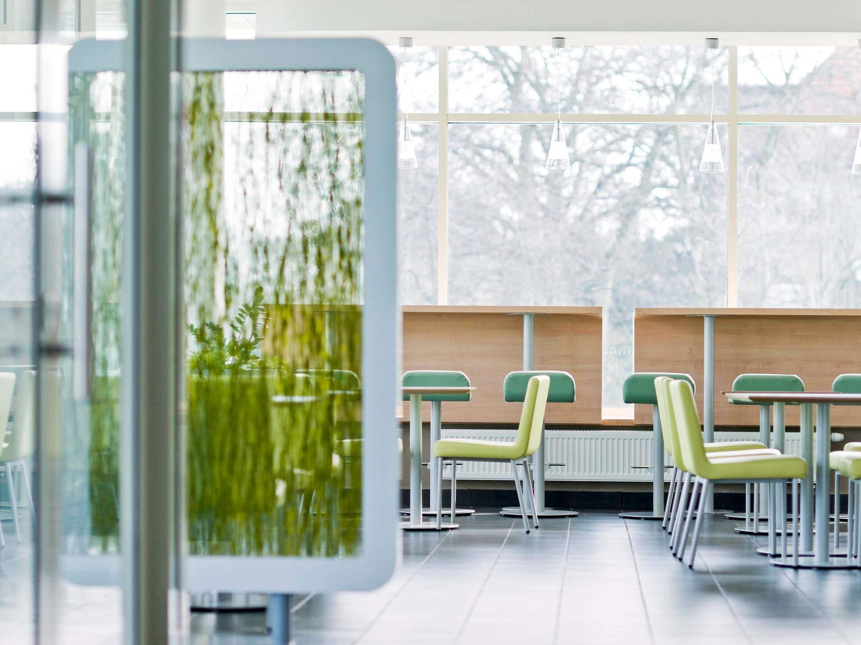 Un espace intérieur moderne, avec une rangée de chaises et de tables vertes, de grandes fenêtres donnant sur des arbres et une cloison en verre agrémentée d’une plante verte.