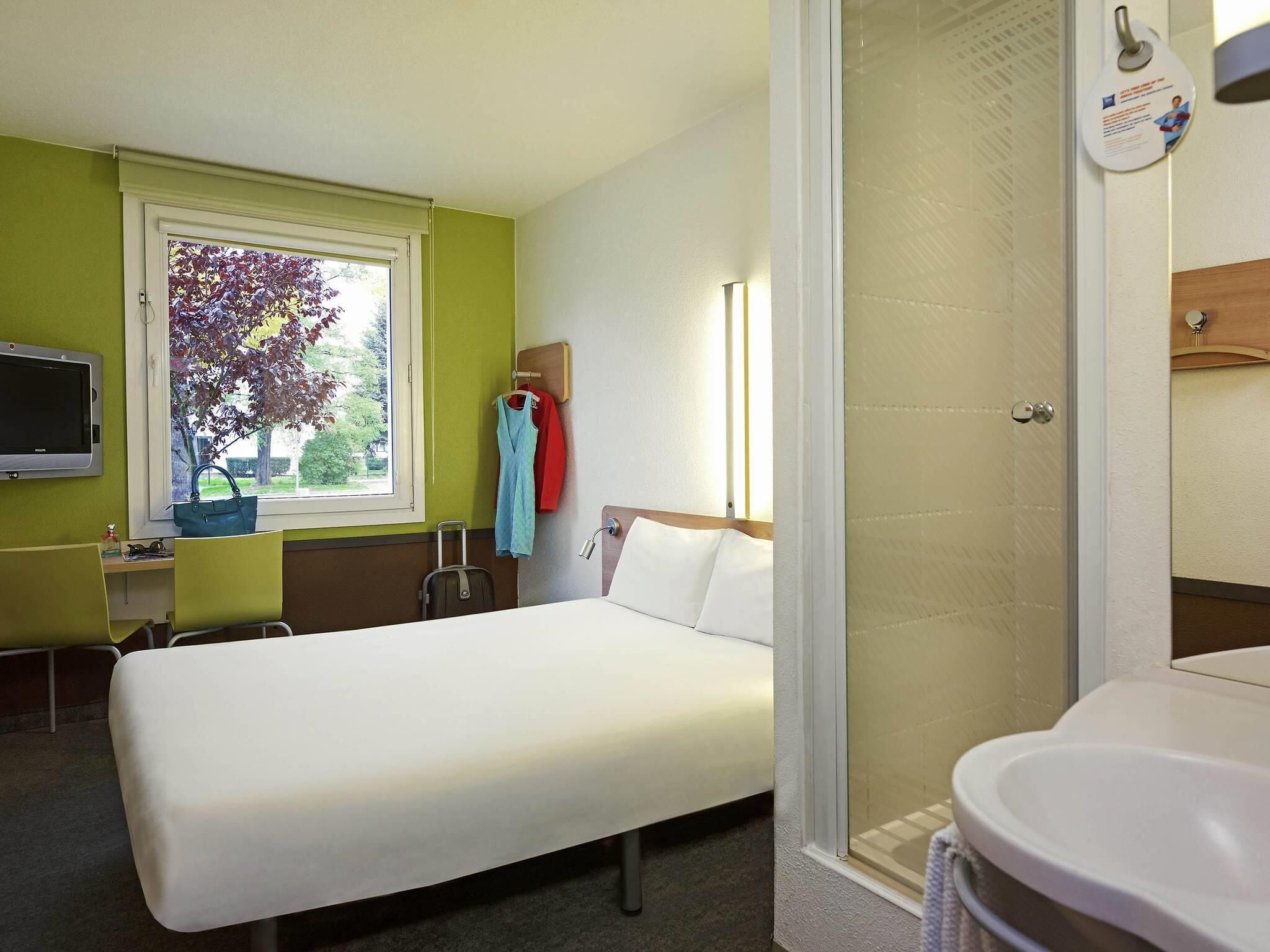 Ein Hotelzimmer mit Bett, Schreibtisch, Stuhl, Fernseher, einem Fenster mit Blick auf Bäume und einem Badezimmer mit Waschbecken und Spiegel.