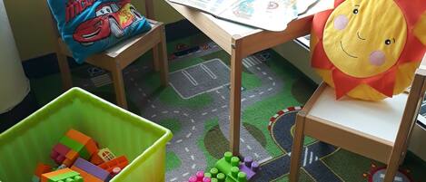 Zona de juegos infantil con un bote verde, una mesa con un libro y una ventana que da a muebles de exterior.