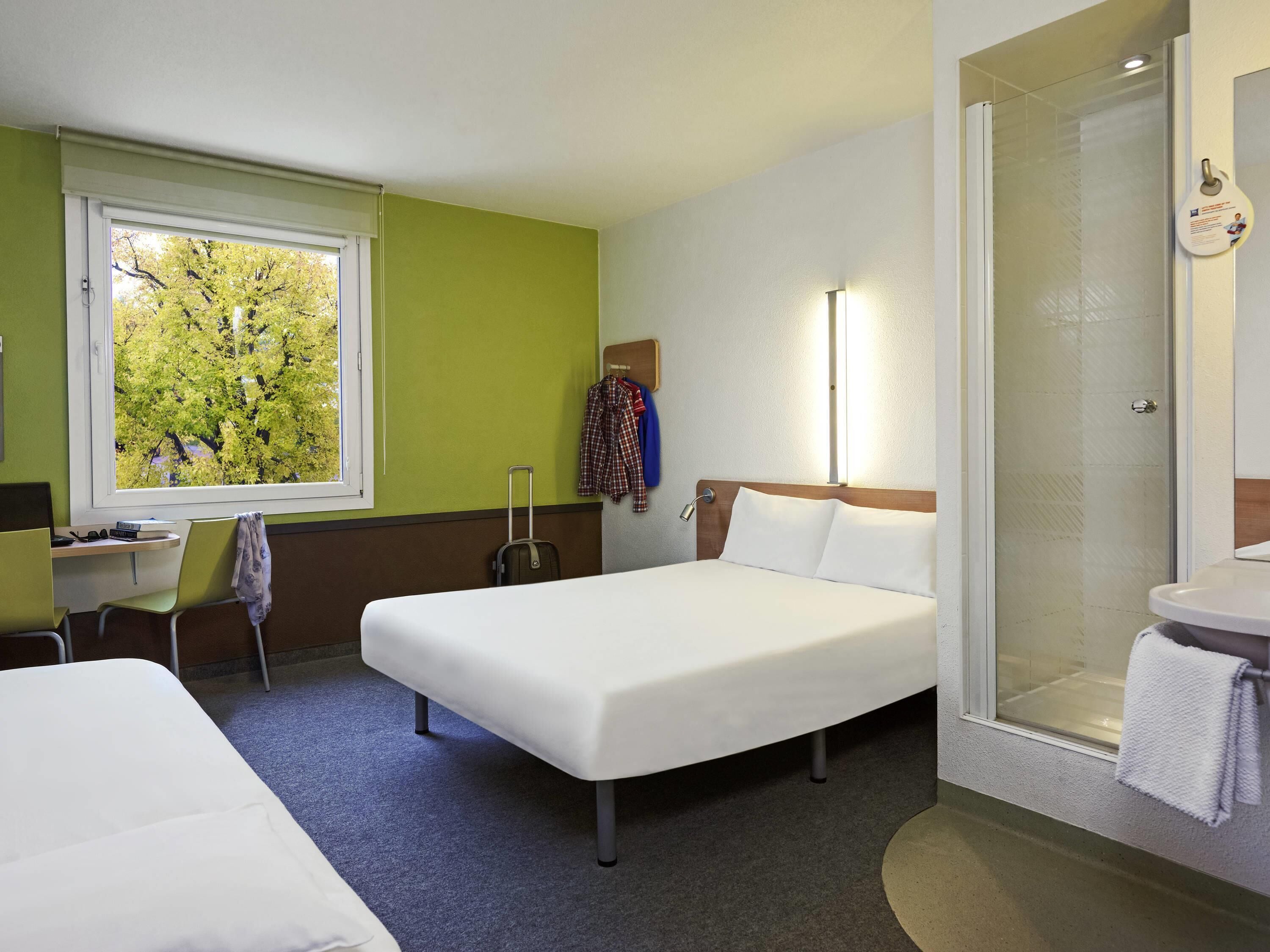 Ein Hotelzimmer mit Bett, Schreibtisch, Stuhl, Koffer, Dusche und einem Fenster mit Blick auf Bäume.