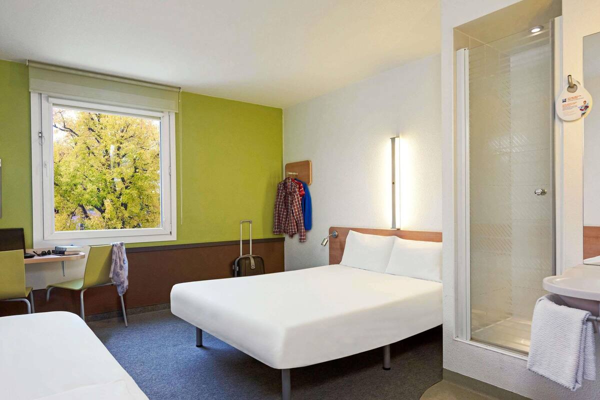 A hotel room with two beds, a desk, a chair, a suitcase, a shower, and a window with a view of trees.