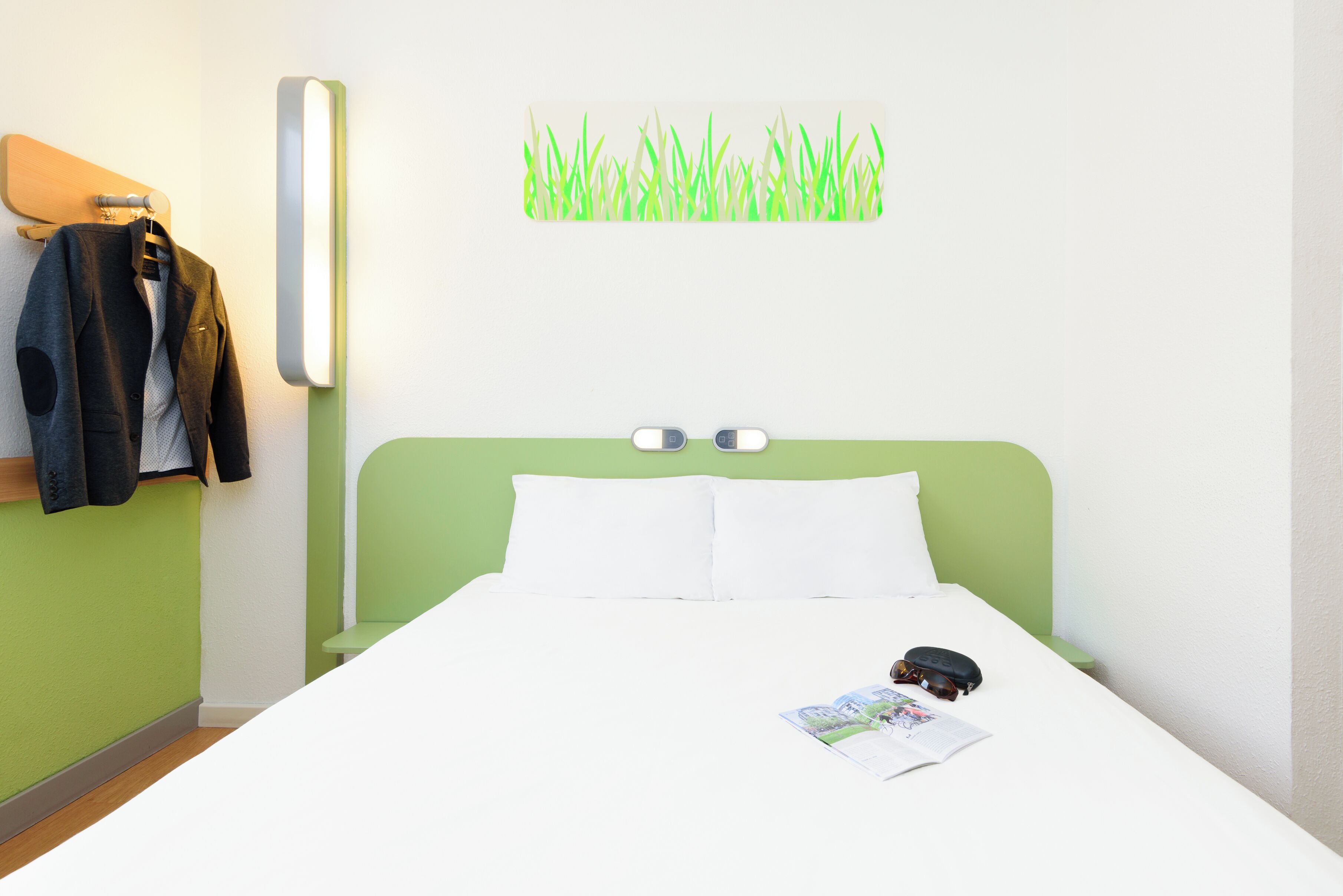 A neatly made bed with white linens, a green headboard, and a wall-mounted light fixture.