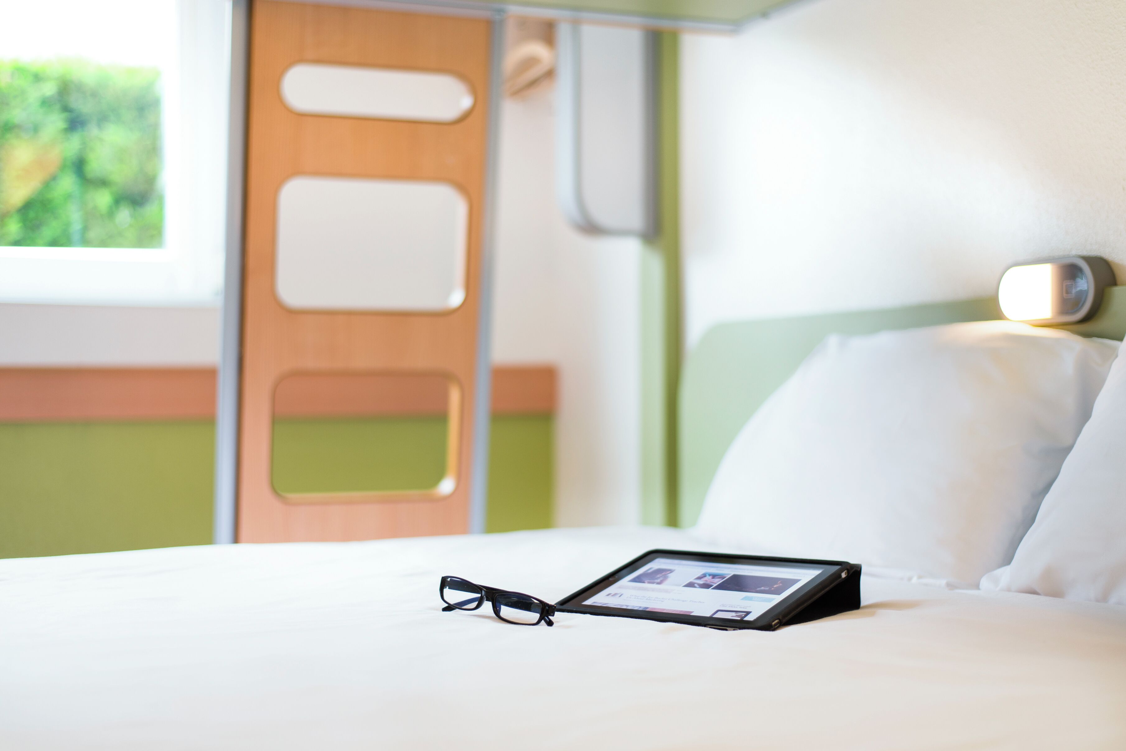 Una cama bien hecha con una tableta y gafas encima, una puerta de madera y una pared verde.