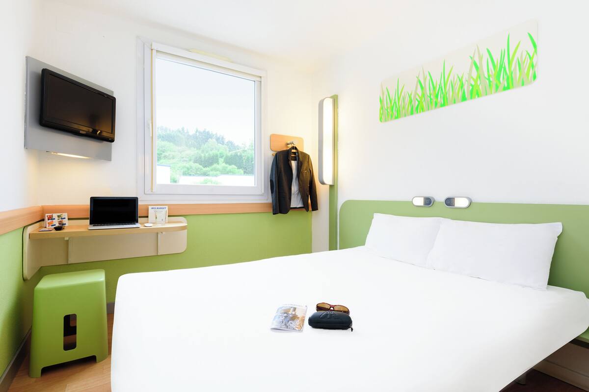 Een hotelkamer met een bed, een bureau met een laptop, een groene kruk en een aan de muur bevestigde televisie.