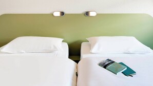 Camera d'albergo con due letti, entrambi con biancheria e cuscini bianchi. Sopra i letti, una lampada a parete e, sopra la testiera, un piccolo quadro raffigurante un prato.