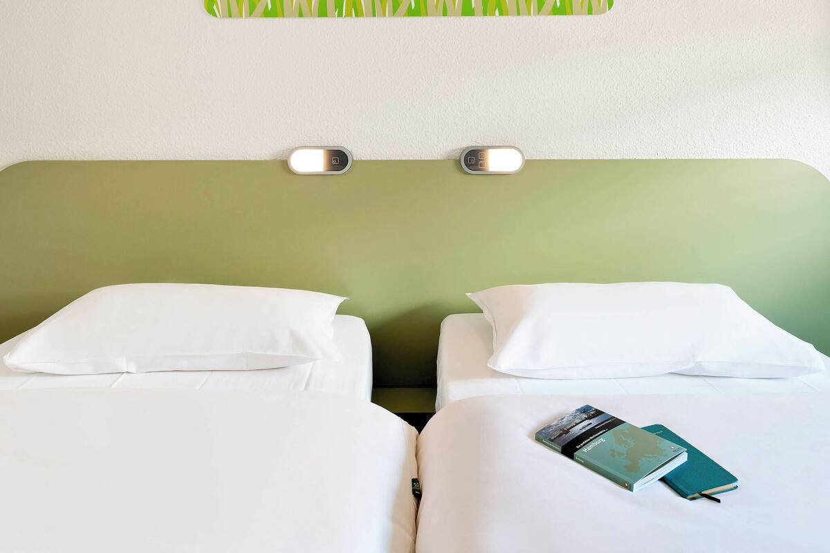 Hotelkamer met twee bedden, elk voorzien van witte beddengoed en kussens. Daarboven is een wandlamp en een klein schilderij van gras boven het hoofdeinde.