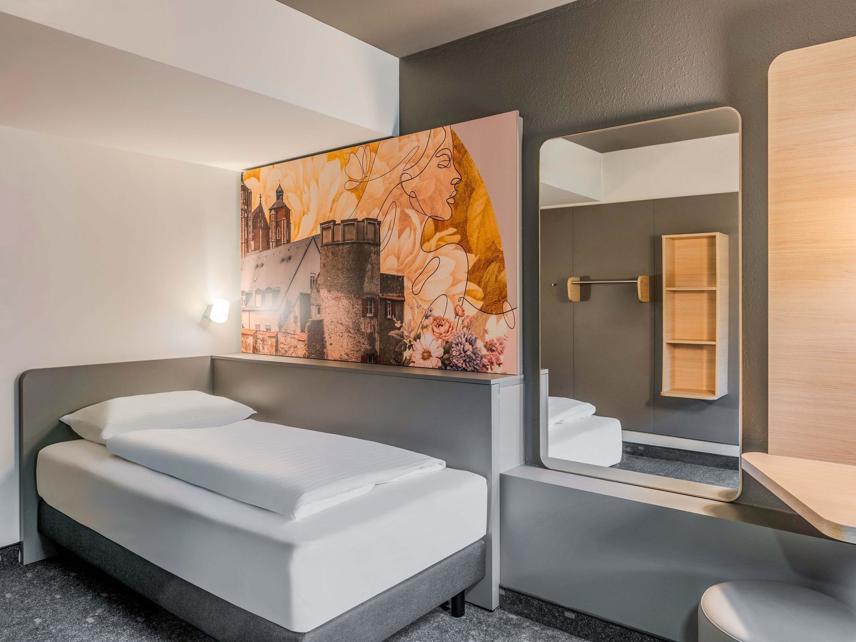 A modern hotel room with a bed, a large mirror, a wooden shelf, and a wall-mounted artwork depicting a cityscape.