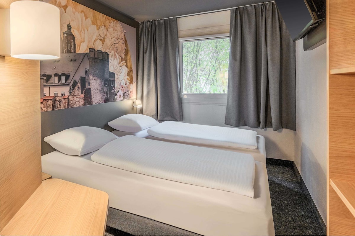 A hotel room with two beds, a window with curtains, a wall-mounted TV, and a painting of a building.