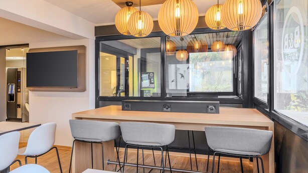 A modern bar area with a wooden counter, four grey upholstered bar stools, and three pendant lights with a ribbed design. A large mirror reflects the interior, and a television screen is mounted on the wall. - B&B HOTEL Ingolstadt-Lenting (Lenting)