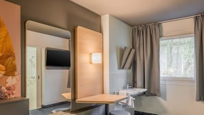 A modern bathroom with a large mirror, a sink, a toilet, and a shower. There is a window with curtains and a view of trees. - B&B HOTEL Ingolstadt-Lenting (Lenting)
