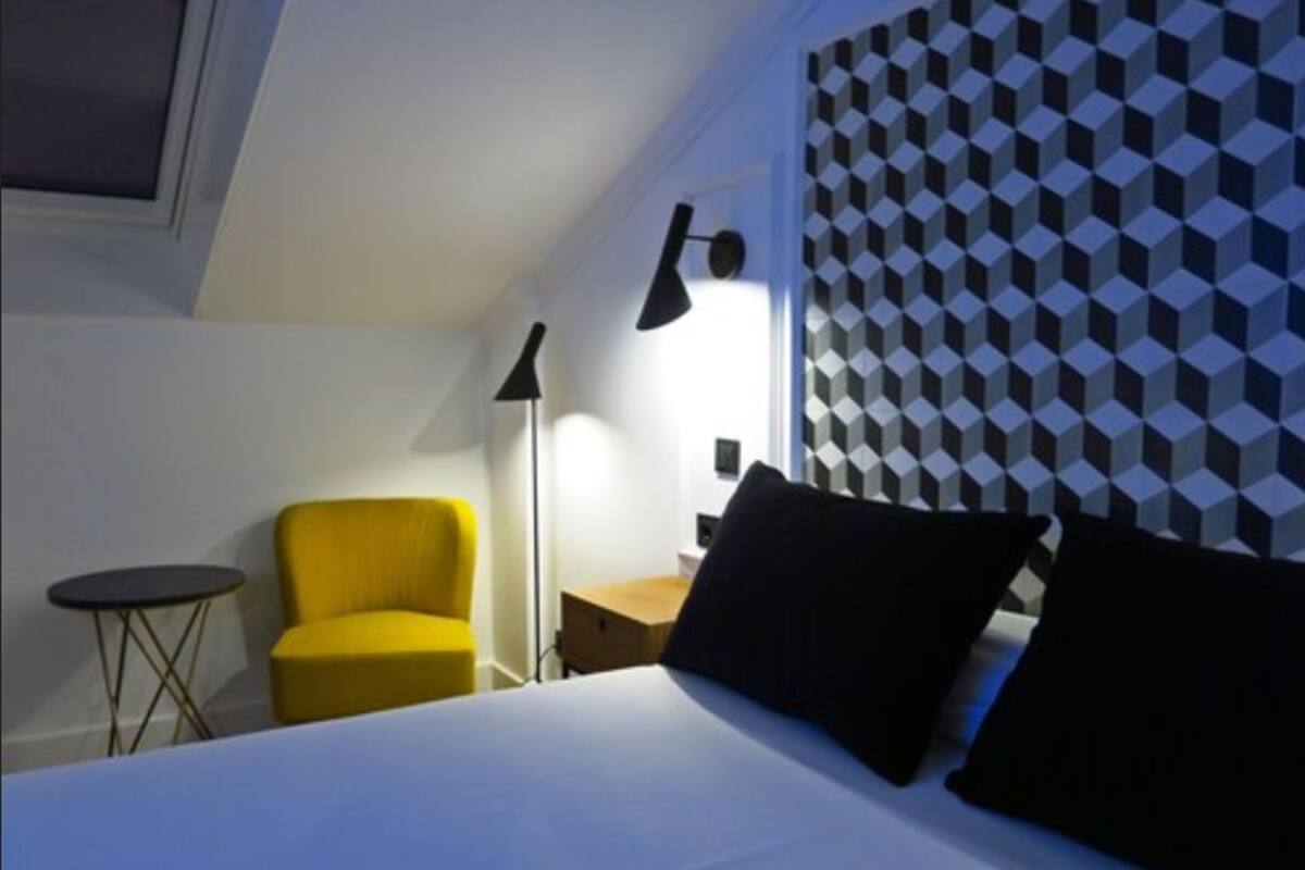 Un dormitorio moderno con una cama blanca, un sillón amarillo, una mesita redonda pequeña y una pared con estampado geométrico.
