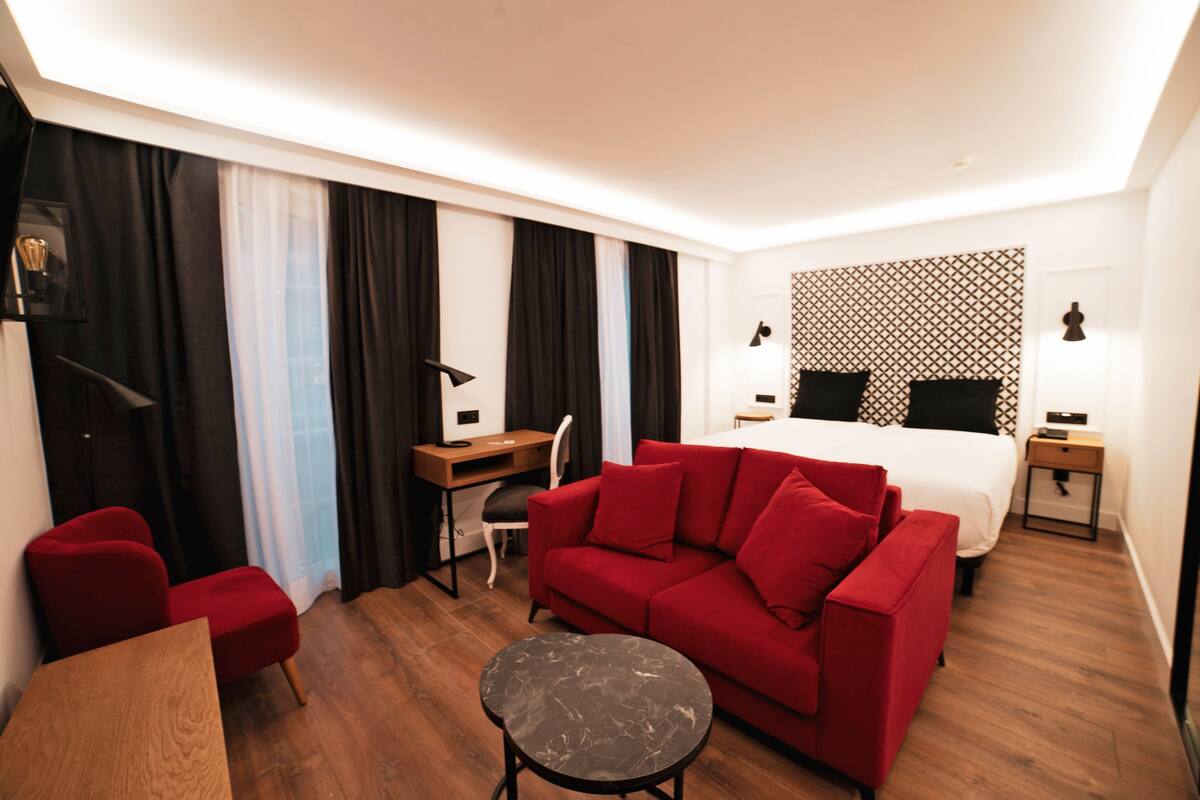 Una habitación de hotel con un sofá rojo, una mesa de madera, una mesita redonda pequeña, un escritorio con una silla, una cama con cabecera estampada y dos mesitas de noche con lámparas.