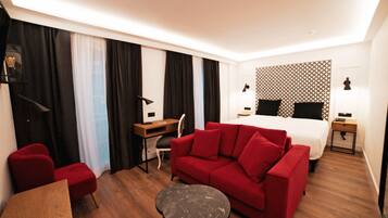 Una habitación de hotel con un sofá rojo, una mesa de madera, una mesita redonda pequeña, un escritorio con una silla, una cama con cabecera estampada y dos mesitas de noche con lámparas.