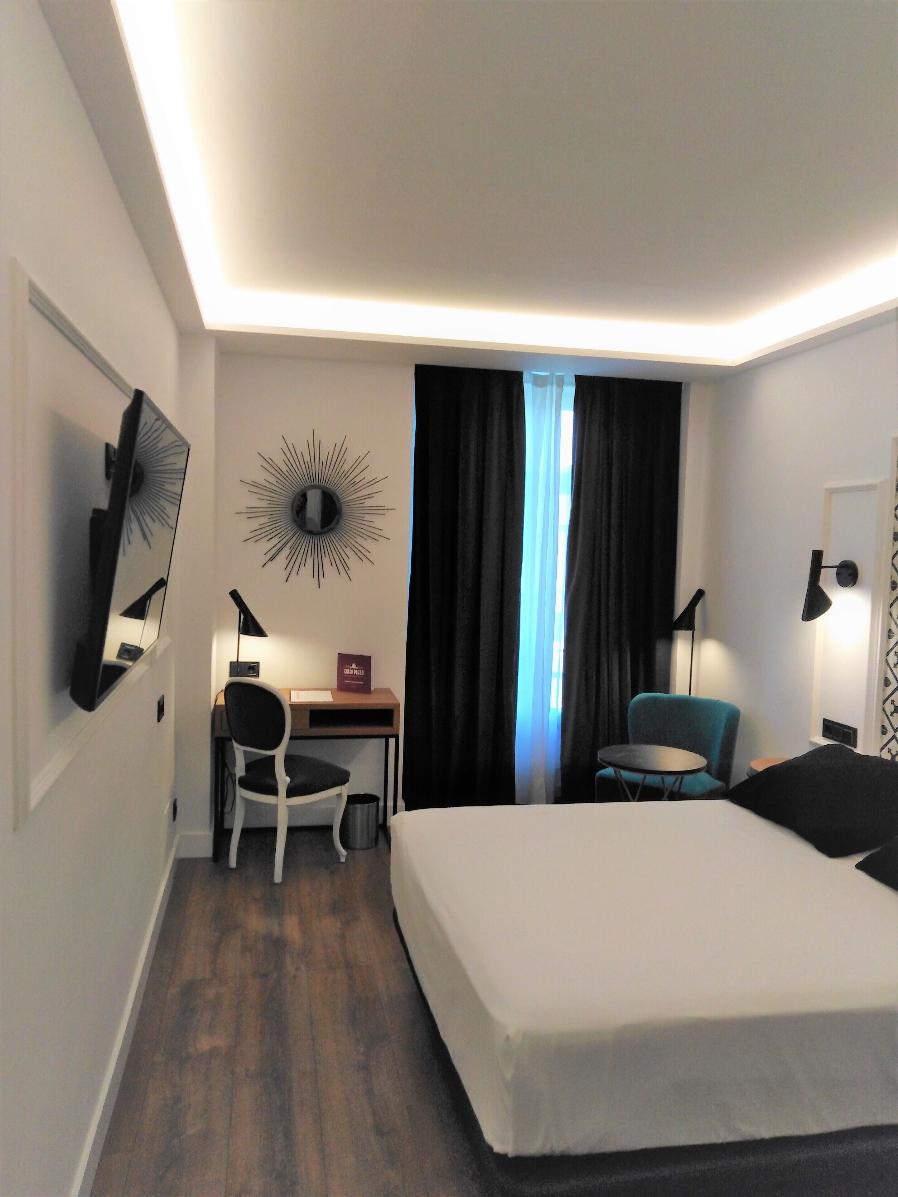 Una habitación de hotel con una cama, un escritorio, una silla, un televisor, un adorno mural con diseño de sol y una ventana con cortinas.