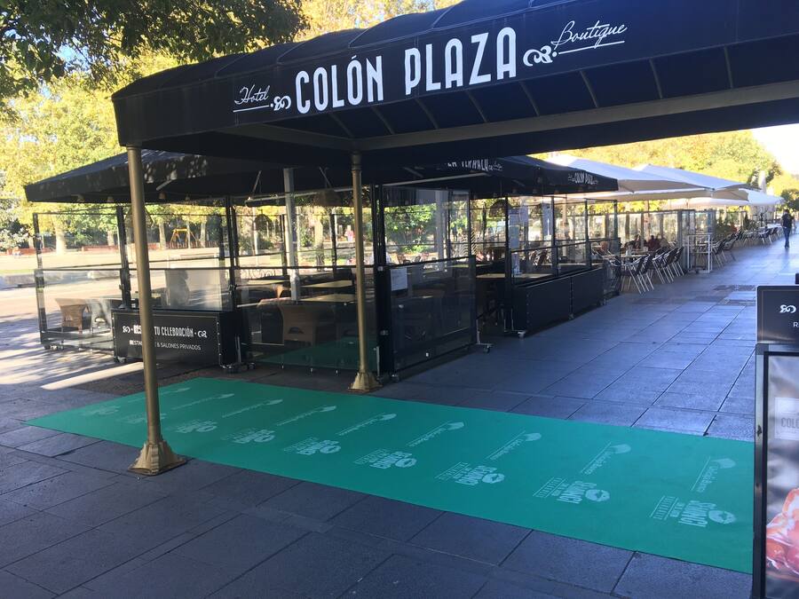 Zona de estar al aire libre cubierta en el Hotel Colón Plaza, con mesas y sillas, un tapete verde con símbolos de reciclaje y una vista despejada del parque circundante.