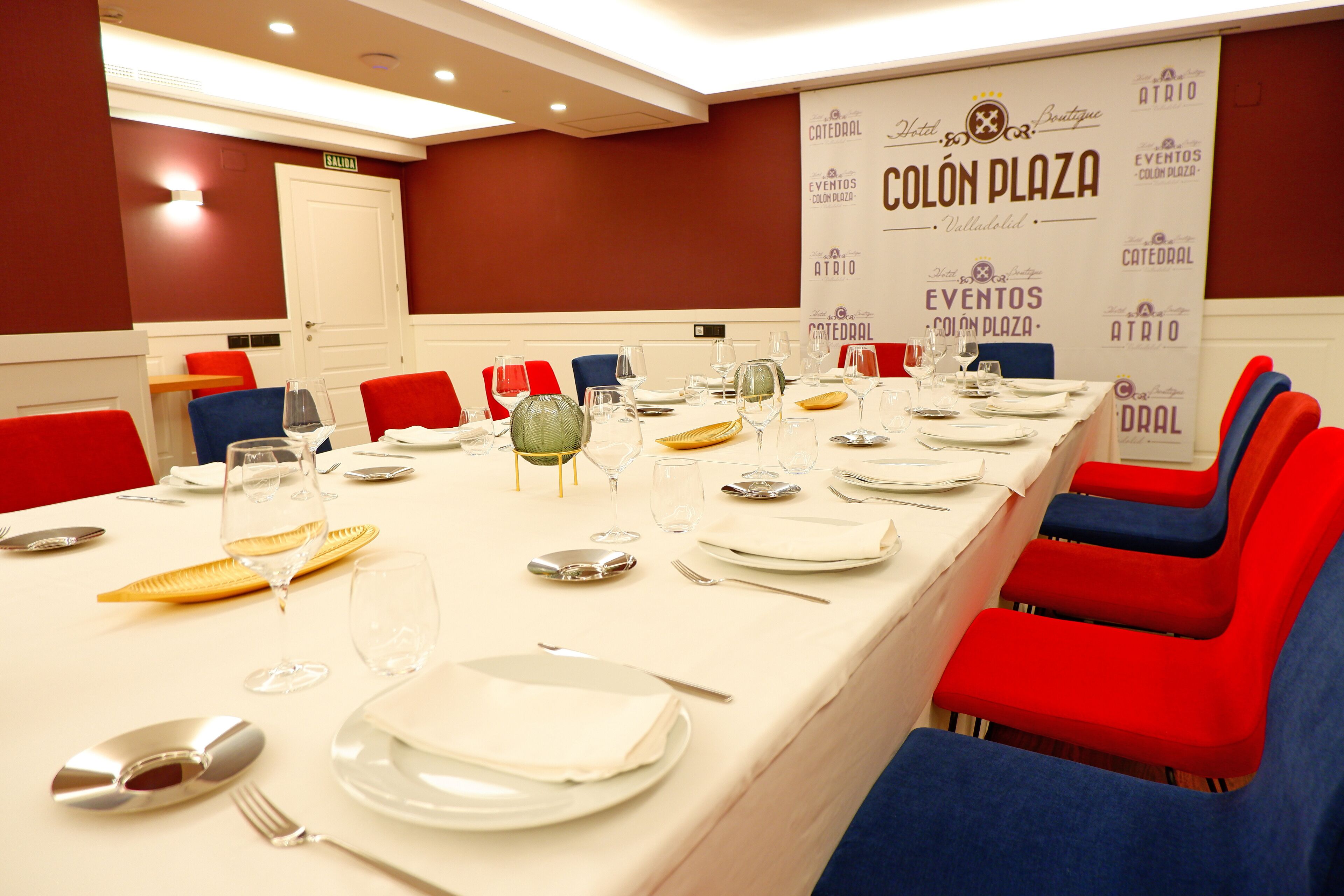 Una sala de conferencias con una larga mesa preparada para una reunión, sillas rojas y azules, y un cartel anunciando eventos.