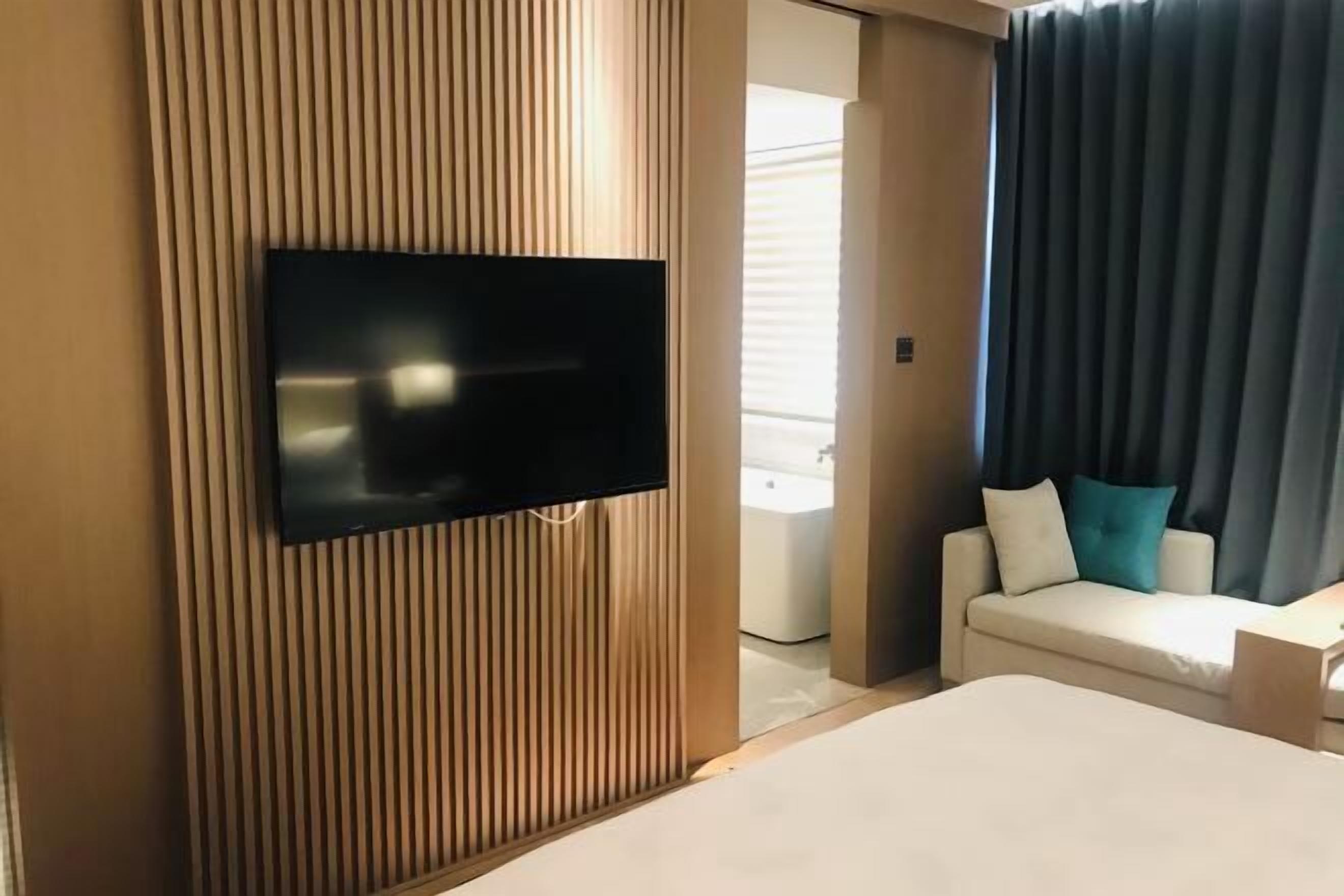 Habitación de hotel con televisor de pantalla plana montado en una pared de paneles de madera, cama con colchón blanco, sofá blanco con cojines turquesa, y baño visible a través de una puerta.