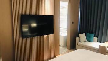 Habitación de hotel con televisor de pantalla plana montado en una pared de paneles de madera, cama con colchón blanco, sofá blanco con cojines turquesa, y baño visible a través de una puerta.