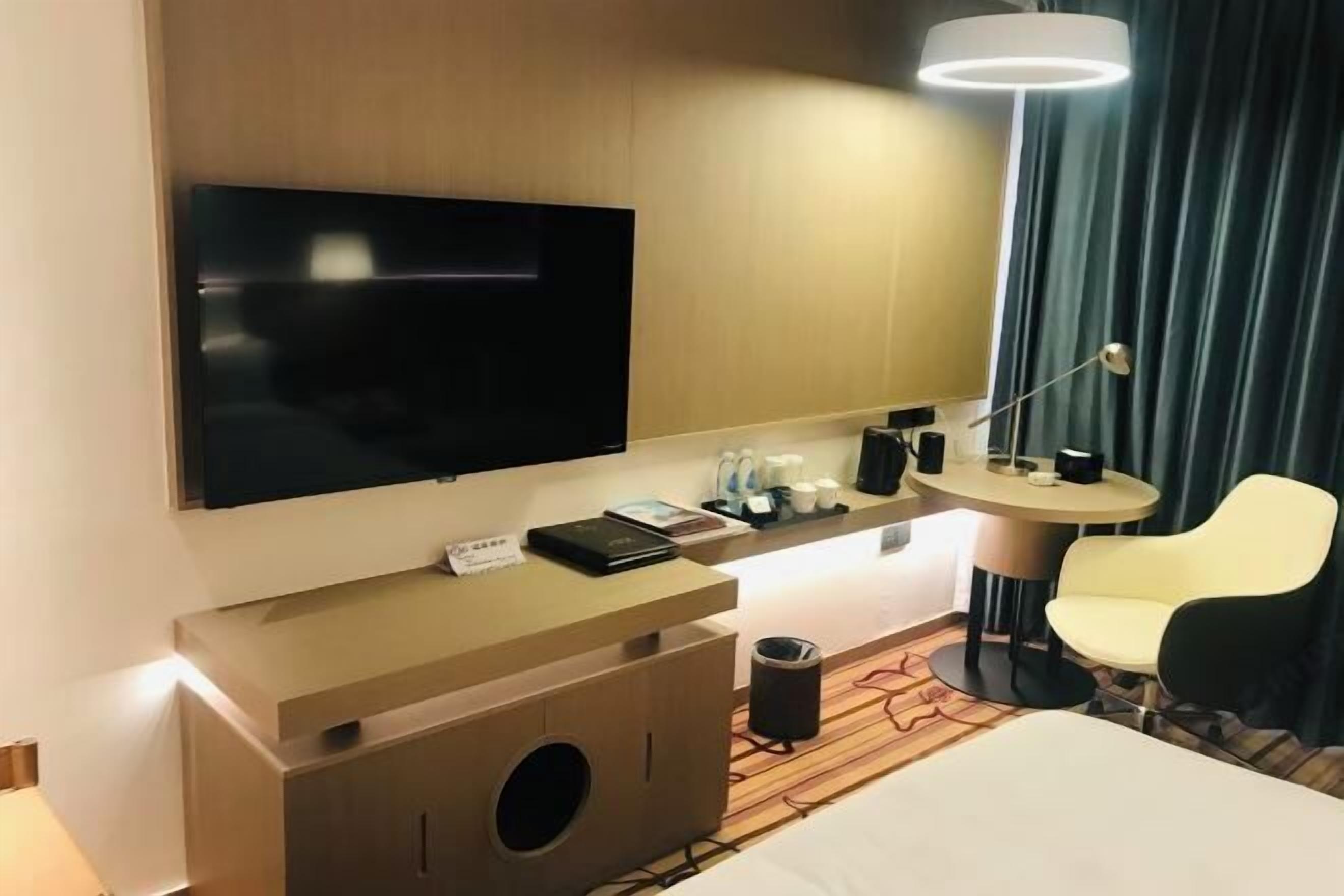A hotel room with a bed, a TV mounted on the wall, a small desk with a lamp, a chair, and a window with curtains.