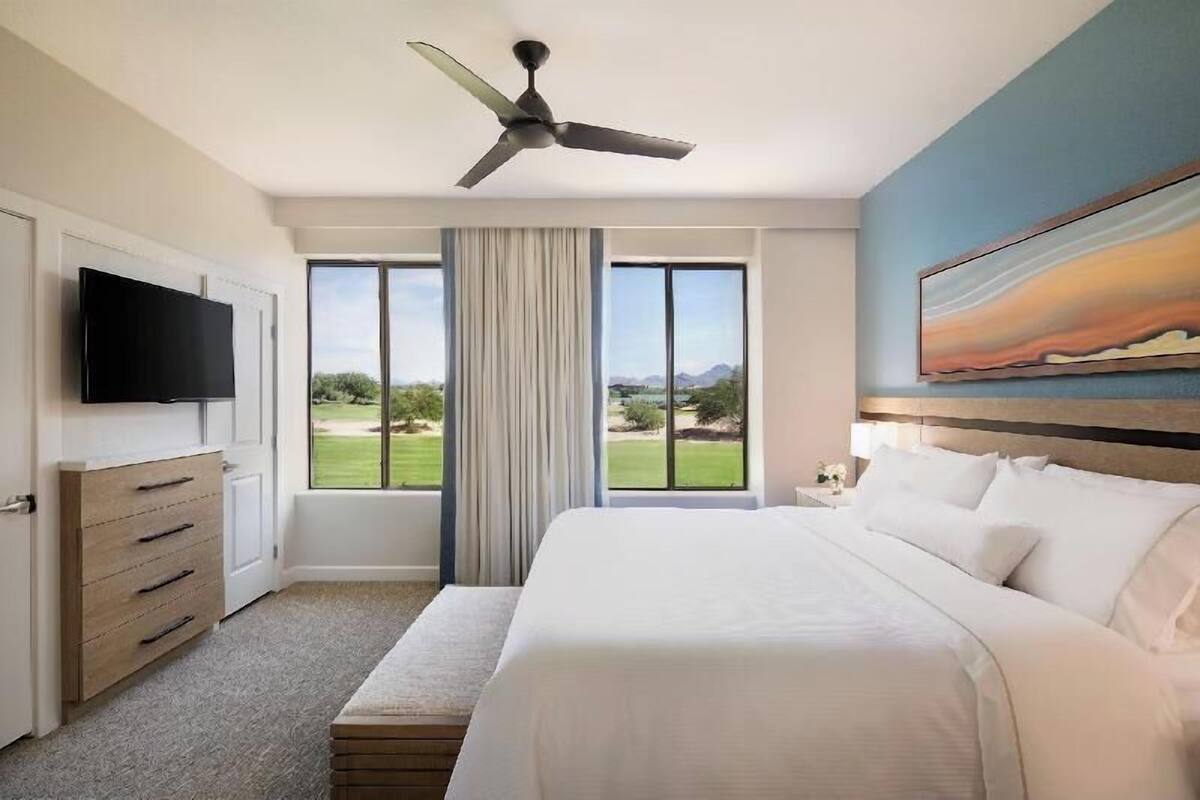 Een hotelkamer met een bed, een plafondventilator, een televisie, een groot raam met uitzicht op de omgeving en een schilderij aan de muur.
