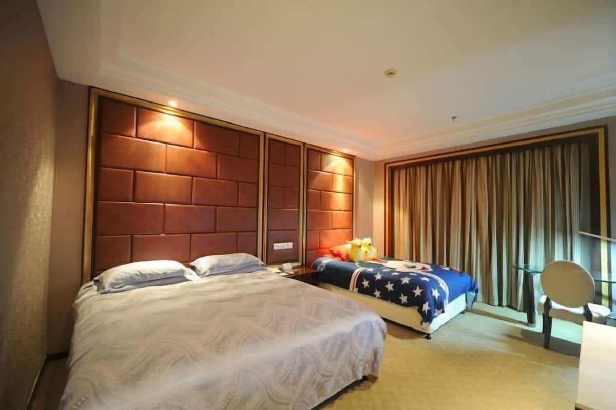 A hotel room with a large bed, a smaller bed, a headboard, a window with curtains, and a bedside table with a mirror.
