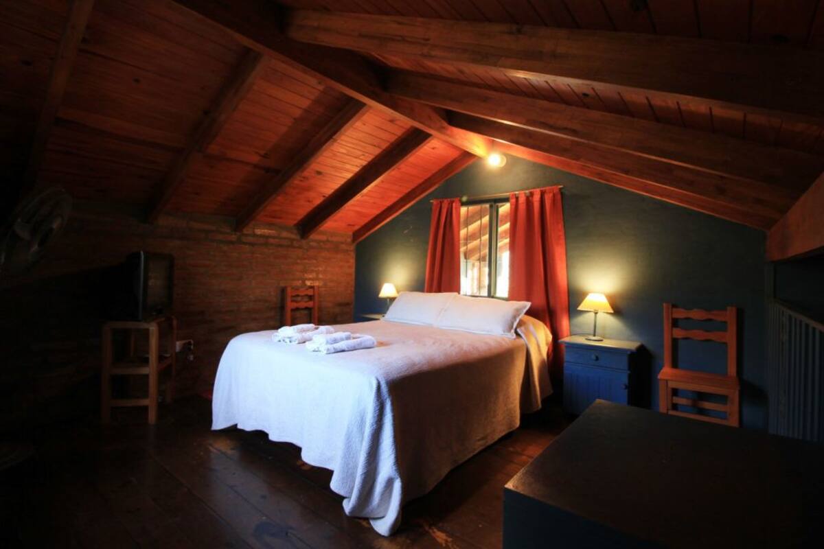 A cozy attic bedroom with a wooden ceiling, a bed with white linens, a nightstand with a lamp, and a chair.