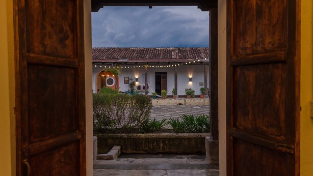 An open wooden door leading to a courtyard with a tiled roof, potted plants, and string lights. - Lemon Tree Hostel (Antigua Guatemala)