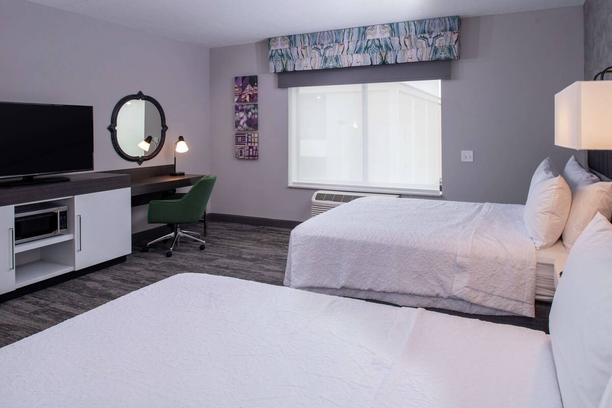 A hotel room with two beds, a desk with a flat-screen TV, a chair, a large window, and a wall-mounted artwork.