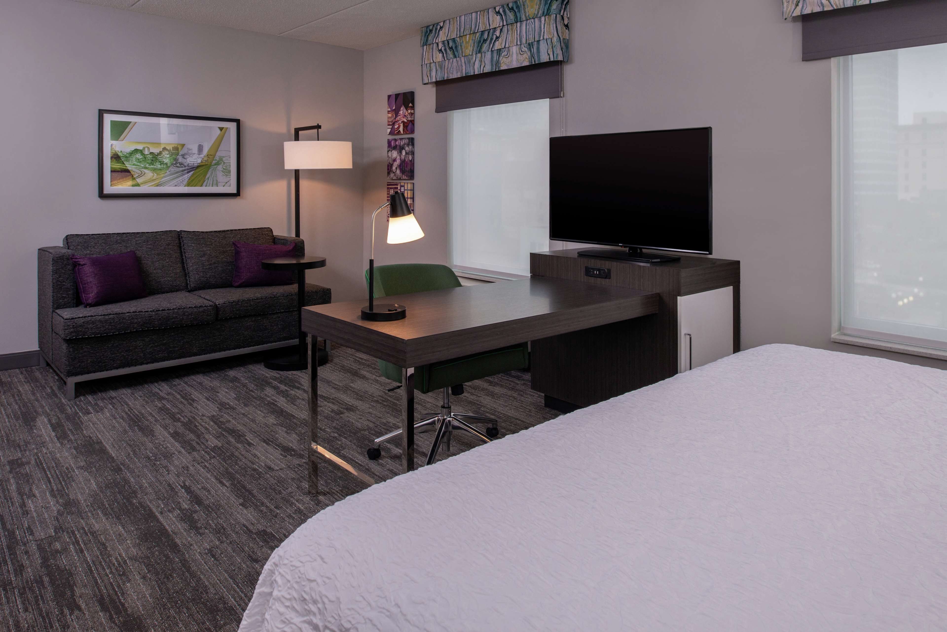Habitación de hotel con una cama, un escritorio con lámpara, un televisor, un sofá con cojines morados y obras de arte en las paredes.
