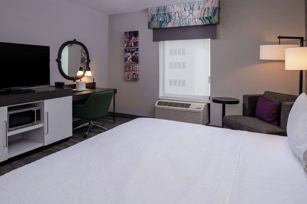 A hotel room with a bed, a desk, a flat-screen TV, a mirror, a window with blinds, a wall-mounted air conditioner, and a lamp.